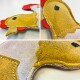 Eine Nahaufnahme eines plüschigen, hühnerförmigen Hundespielzeugs, das gelbes und rotes Filz mit gestickten Details und einem lächelnden Gesicht zeigt.