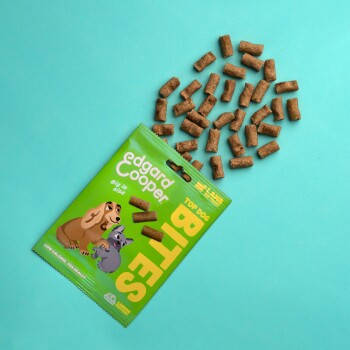 Grüne Verpackung von Edgard Cooper Top Dog Bites mit Cartoon-Hunden, umgeben von kleinen Hundeleckerlis auf einem türkisfarbenen Hintergrund.