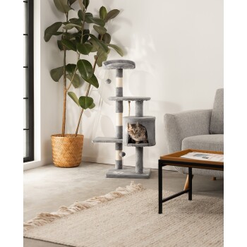 Ein gemütliches Wohnzimmer mit einem grauen Katzenbaum, aus dem eine getigerte Katze aus einem Fach schaut, einem geflochtenen Pflanzenhalter und einem Couchtisch.
