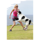 Eine Frau in einem pinken Sportoutfit spielt mit einem springenden schwarz-weißen Hund auf einem grünen Feld, mit dem Ozean im Hintergrund.