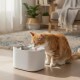 Orangegetigerte Katze trinkt aus weißem ASTROPET-Trinkbrunnen mit silberner Tülle im Wohnzimmer.
