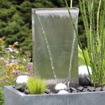 Waterfall Kit 30 Ein moderner Wasserbrunnen mit einer eleganten, transparenten Platte, aus der Wasser herabfließt, umgeben von dekorativen silbernen Kugeln und grünen Pflanzen.