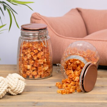 Zwei Glasbehälter gefüllt mit orangefarbenen Haustierleckereien, ein Behälter umgekippt und Leckereien auf einem Holztisch verschüttet, mit einem gemütlichen Haustierbett im Hintergrund.