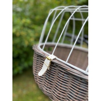 Eine Nahaufnahme eines Korb-Haustierträgers mit einem weißen Metallrahmen und einem goldenen Schnallenriemen, vor einem verschwommenen grünen Hintergrund.