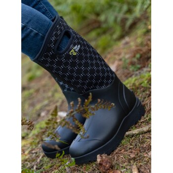 Schwarze wasserdichte Stiefel mit einem gemusterten Obermaterial, getragen von einer Person, die auf dem Waldboden mit Grün und Zweigen steht.