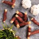 Eine schwarze Schüssel, gefüllt mit Hundeleckerlis, speziell gerollten Sticks, die in Fleisch gewickelt sind, umgeben von weiteren Leckerlis und zwei Seilspielzeugen auf einer grauen Oberfläche.