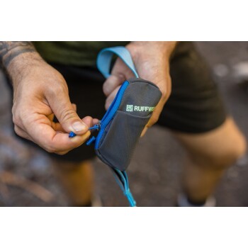 Eine Person hält einen grauen und blauen Hundekotbeutelspender mit dem "RUFFWEAR"-Logo und öffnet ihn, um auf die Beutel im Inneren zuzugreifen.
