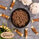 Draufsicht auf eine schwarze Schüssel mit Nassfutter, umgeben von mit Fleisch umwickelten Kauknochen, Tau-Spielzeugen und dem 'Gino's'-Logo.