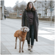 Eine Frau steht auf einer Stadtstraße mit einem grauen Weimaraner-Hund, der einen braunen Mantel trägt. Die Frau ist in einem langen schwarzen Mantel und lässiger Kleidung gekleidet.