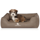 Ein kleiner, flauschiger Welpe mit braunem und weißem Fell sitzt in einem gemütlichen, taupefarbenen Hundebett mit dem Logo "KNUFFELWUFF".