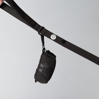 Eine schwarze Haustierleine mit einem kleinen angehängten Beutel mit floralem Design, gehalten von einer Hand vor einem hellgrauen Hintergrund.