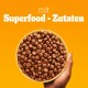 Eine Hand hält eine gelbe Schüssel, gefüllt mit kleinen, runden Haustierfutterkroketten, vor einem hellorangefarbenen Hintergrund, mit dem Text "mit Superfood - Zutaten."