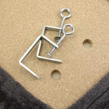 Metallteile zur Montage eines Haustierbettes, einschließlich einer U-förmigen Halterung und einer Schraube, die auf einer Holzoberfläche mit einem weichen grauen Stoffrand liegen.