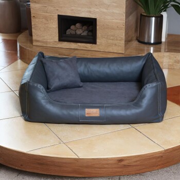 Schwarzes Lederhaustierbett mit Kissen und Etikett „tierlandade“ auf gefliestem Boden nahe Kamin und Pflanze.