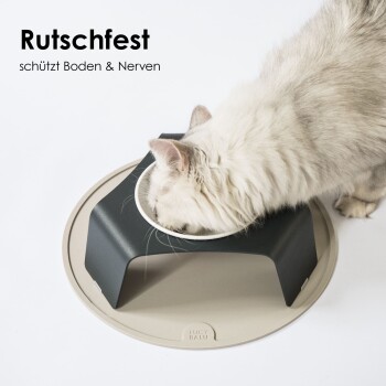 Eine flauschige weiße Katze, die aus einer modernen erhöhten Schüssel mit schwarzem Boden und beigem Tablett frisst, auf dem steht: "Rutschfest schützt Boden & Nerven."