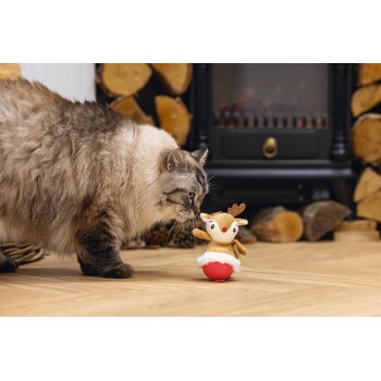 Fluffy cat sniffing a small reindeer plush toy with a red base on a wooden floor near stacked firewood.
