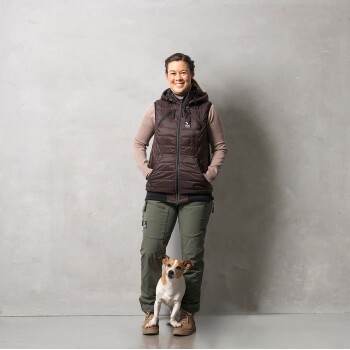 Eine Frau in einer braunen Daunenweste und grünen Hosen steht mit ihrem kleinen Hund, einem Jack Russell Terrier, vor einem grauen Hintergrund.