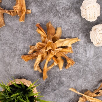 Ein Haufen getrockneter Tierkausnacks auf einer grauen Oberfläche, umgeben von einer kleinen grünen Pflanze in einem geflochtenen Korb und zwei weißen Seilspielzeugen.