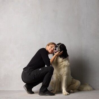 Eine Frau in schwarzer Kleidung kniet neben einem großen, flauschigen Hund und berührt sanft sein Gesicht, während der Hund zu ihr aufblickt.