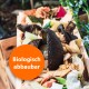 Eine Holzkiste gefüllt mit verschiedenen organischen Abfällen, einschließlich Avocado-Schalen und Gemüseabfällen, mit einem orangefarbenen Kreis, der "Biologisch abbaubar" besagt.