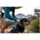 Eine Frau in einem türkisfarbenen Langarmshirt befestigt ein blaues Geschirr an einem Rottweiler in einer bergigen Umgebung im Freien.