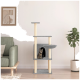 Ein gemütliches Wohnzimmer mit einem modernen Katzenbaum in grauer Ausführung, einer Tabbykatze, die aus einem Fach schaut, und einem grünen Sofa.