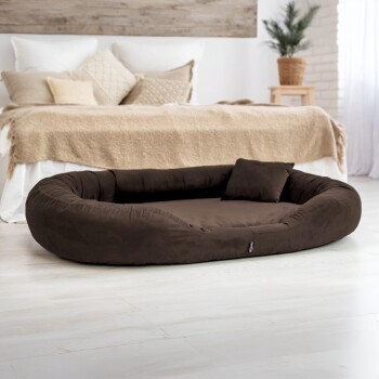 Großes ovales dunkelbraunes Haustierbett mit kleinem Kissen, auf hellem Boden in gemütlichem Schlafzimmer.